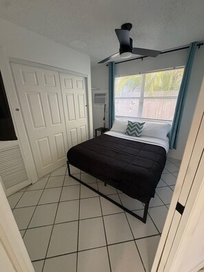 Ein Schlafzimmer mit Bett, Deckenventilator, Fenster mit Jalousien und einer weißen Tür.