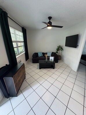 Ein Wohnzimmer mit Deckenventilator, einem schwarzen Sofa, einem Couchtisch, einem an der Wand befestigten Fernseher und einem Fenster mit Vorhängen.