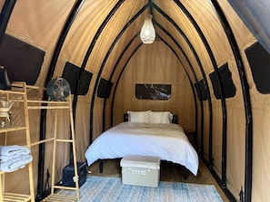 Ein Zelt mit einem Bett, einem Ventilator, einem Regal mit Handtüchern und einem kleinen Tisch.