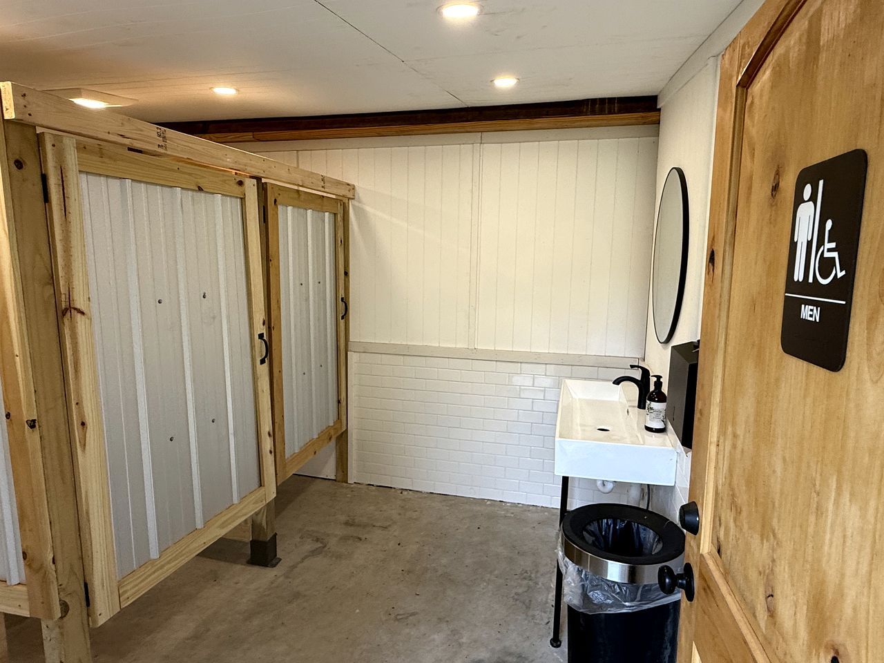 Des toilettes pour hommes avec une porte en bois, un lavabo blanc et une poubelle.