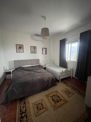 A bedroom with a large bed, a smaller bed, a nightstand, a lamp, a window with curtains, and a wall with three framed pictures.