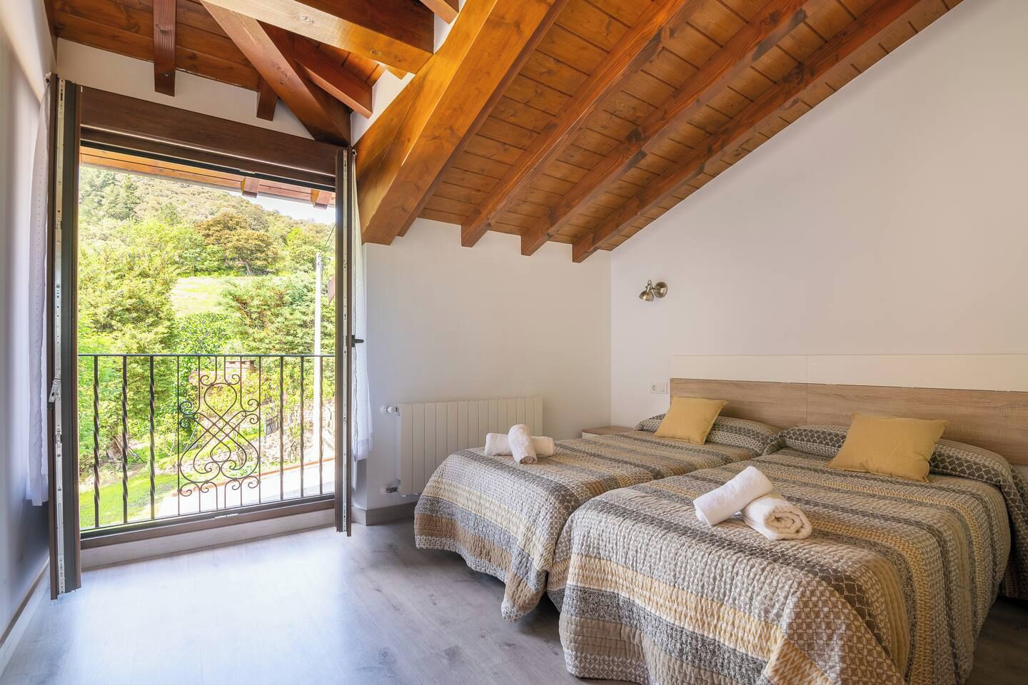 Un dormitorio con dos camas, techo de madera, vista a follaje exterior y balcón con barandilla metálica.