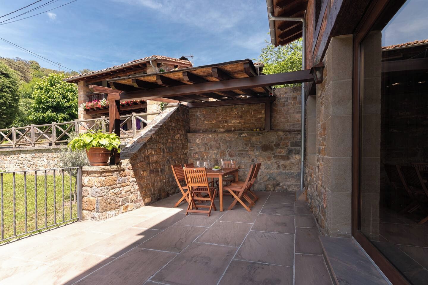 Un patio exterior con paredes de piedra, muebles de madera y una pérgola.