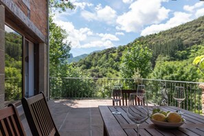 Une table en bois sur laquelle se trouve un saladier rempli de citrons et deux verres à vin, le tout sur un balcon offrant une vue sur la montagne.