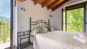 Quarto com cama, mesa de cabeceira, janela com vista para o verde e varanda com grades.