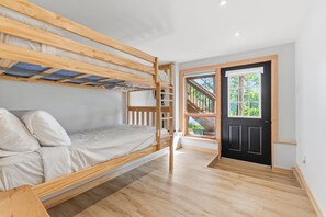 A bunk bed room with a wooden floor, a black door, and a window with a view of greenery. - Lake It Easy | On Portage Lake (Parry Sound District)