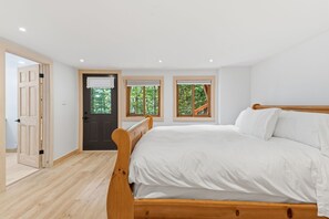 A bedroom with a wooden bed, two windows with blinds, a door, and a view of greenery outside.