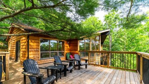 A wooden cabin with a deck, outdoor seating, and a large window overlooking the forest.