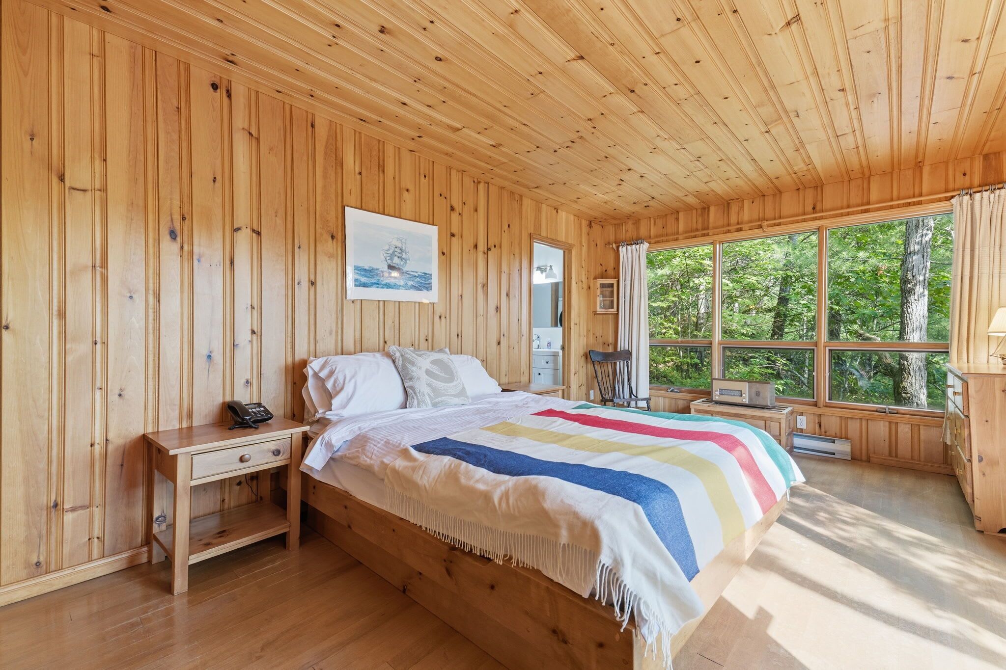 A wooden cabin bedroom with a bed, bedside table, and a window with a view of trees.