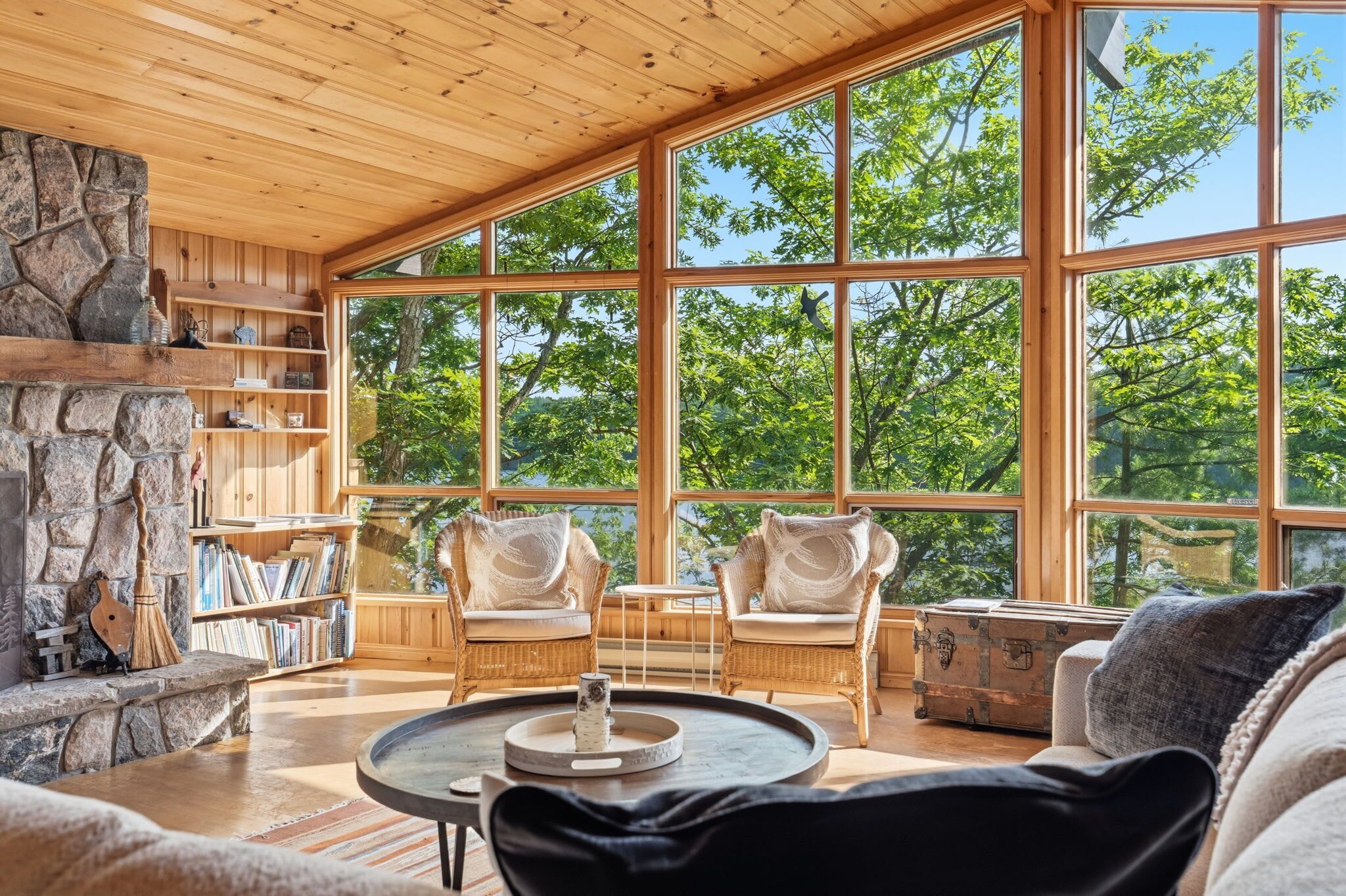 A spacious room with a stone fireplace, wicker furniture, and large windows offering a view of trees.