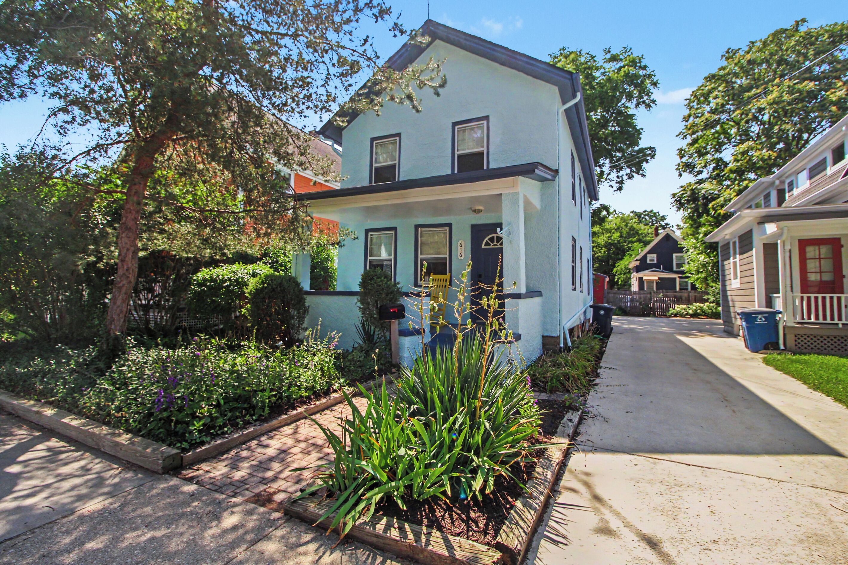 A two-story house with a front porch, a mailbox, and a garden with various plants.