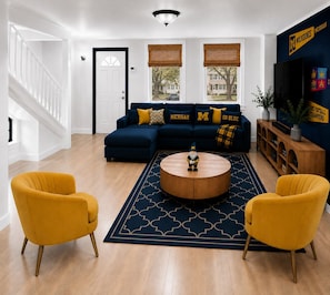 A living room with a dark blue sofa, yellow armchairs, a round coffee table, and a large TV. - #1 Location I 1/2 Mile to Stadium I 1/3 Mile to Downtown I GO BLUE (Ann Arbor)