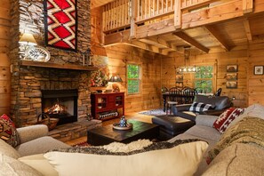 Ein gemütliches Blockhaus-Interieur mit Kamin aus Stein, Holzbalken, Essbereich und Wohnzimmer, möbliert mit Sofas und einem Couchtisch.