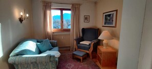 Een gezellige kamer met een gestreept bankje, een donkere fauteuil, een klein tafeltje met een lamp en een raam met gordijnen.