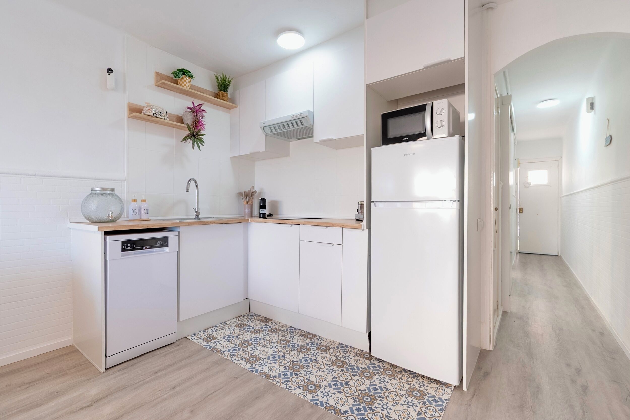 A modern kitchen with white cabinetry, a dishwasher, a microwave, and a refrigerator. There are wooden shelves with decorative items and a patterned rug on the floor.