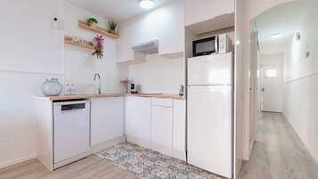 A modern kitchen with white cabinetry, a dishwasher, a microwave, and a refrigerator. There are wooden shelves with decorative items and a patterned rug on the floor.