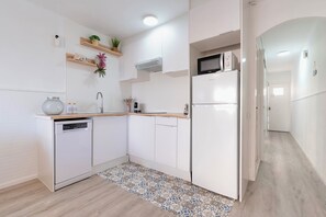 A modern kitchen with white cabinetry, a dishwasher, a microwave, and a refrigerator. There are wooden shelves with decorative items and a patterned rug on the floor. - Beachfront Apartment with Terrace & Ocean Views (Cambrils)