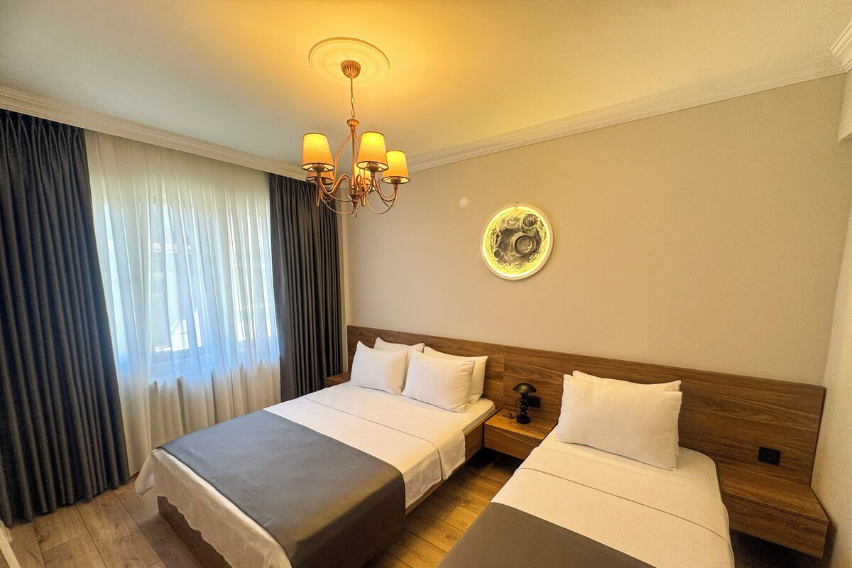Een hotelkamer met een tweepersoonsbed, een nachtkastje met lamp, een wandklok en een raam met gordijnen.