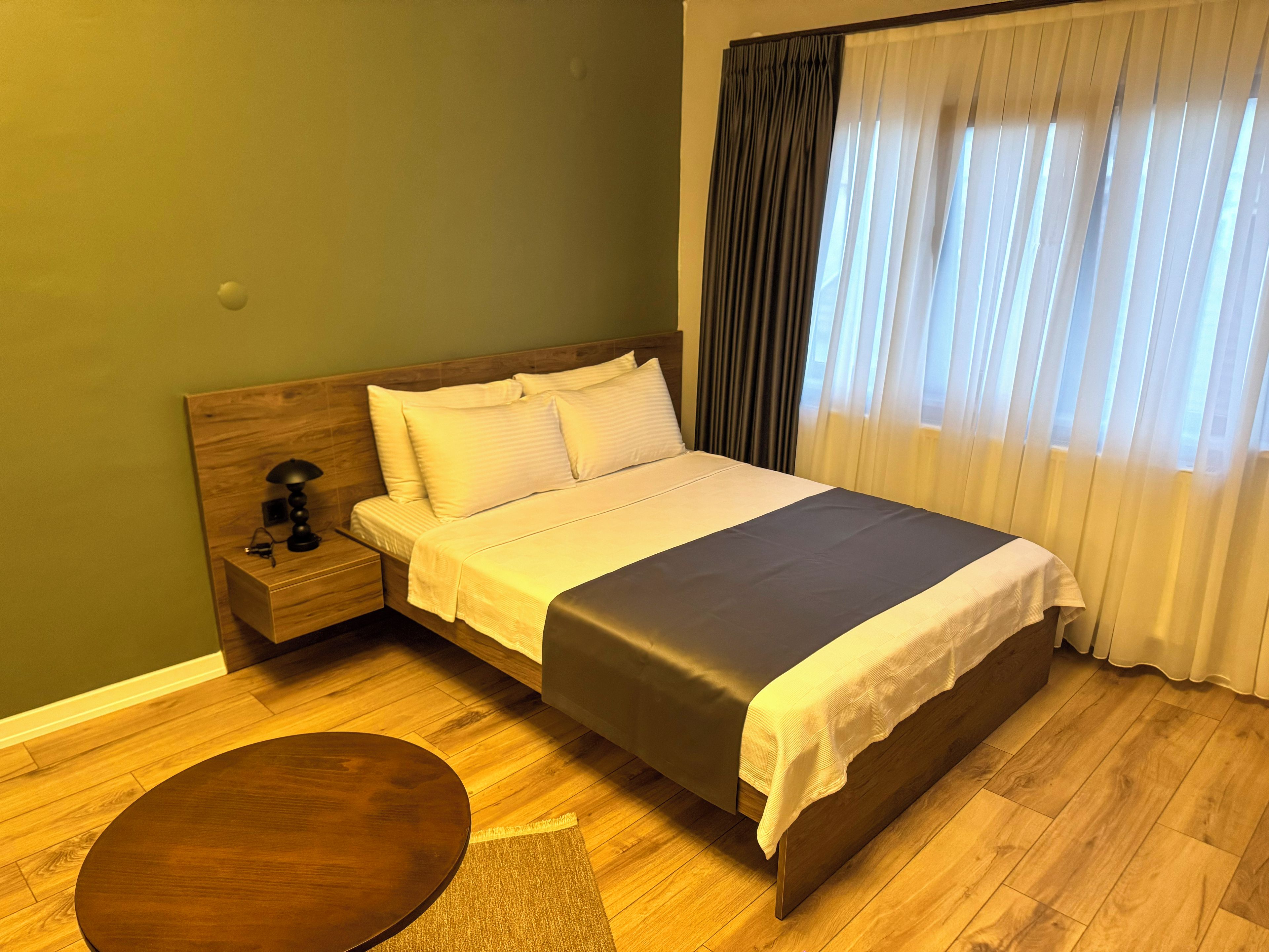 A hotel room with a wooden bed, a bedside table with a lamp, a round wooden table, and a window with curtains.