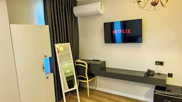 Ein Hotelzimmer mit Flachbildfernseher, der 'NETFLIX' anzeigt, einem Schreibtisch mit Stuhl, einem Kleiderschrank und einer Klimaanlage.