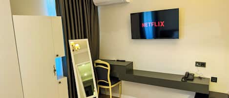 A hotel room with a flat-screen TV displaying 'NETFLIX', a desk with a chair, a wardrobe, and an air conditioning unit.