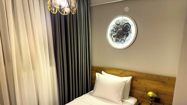 A hotel room with a bed, a nightstand, a lamp, a wall-mounted circular light, and a window with curtains. - Fethibey Otel (Bartın)