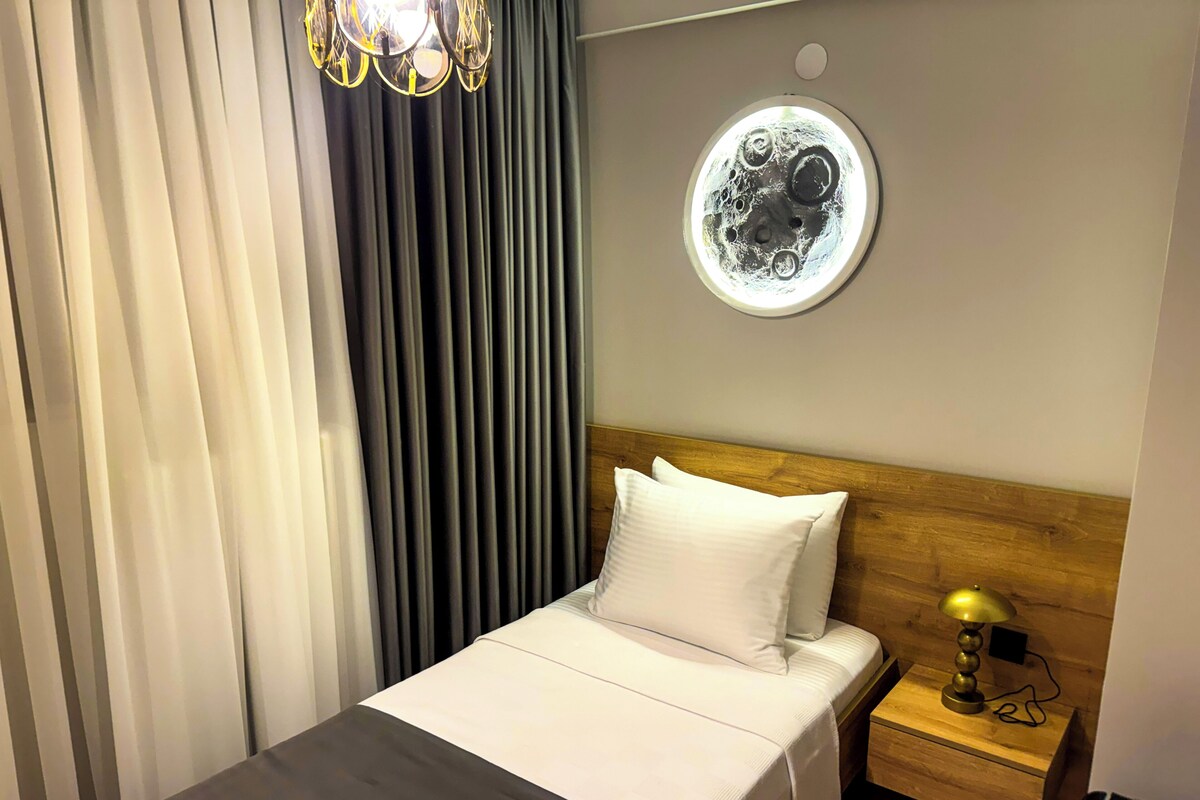Hotelkamer met een bed, nachtkastje, lamp, rond licht aan de muur, en een raam met gordijnen.