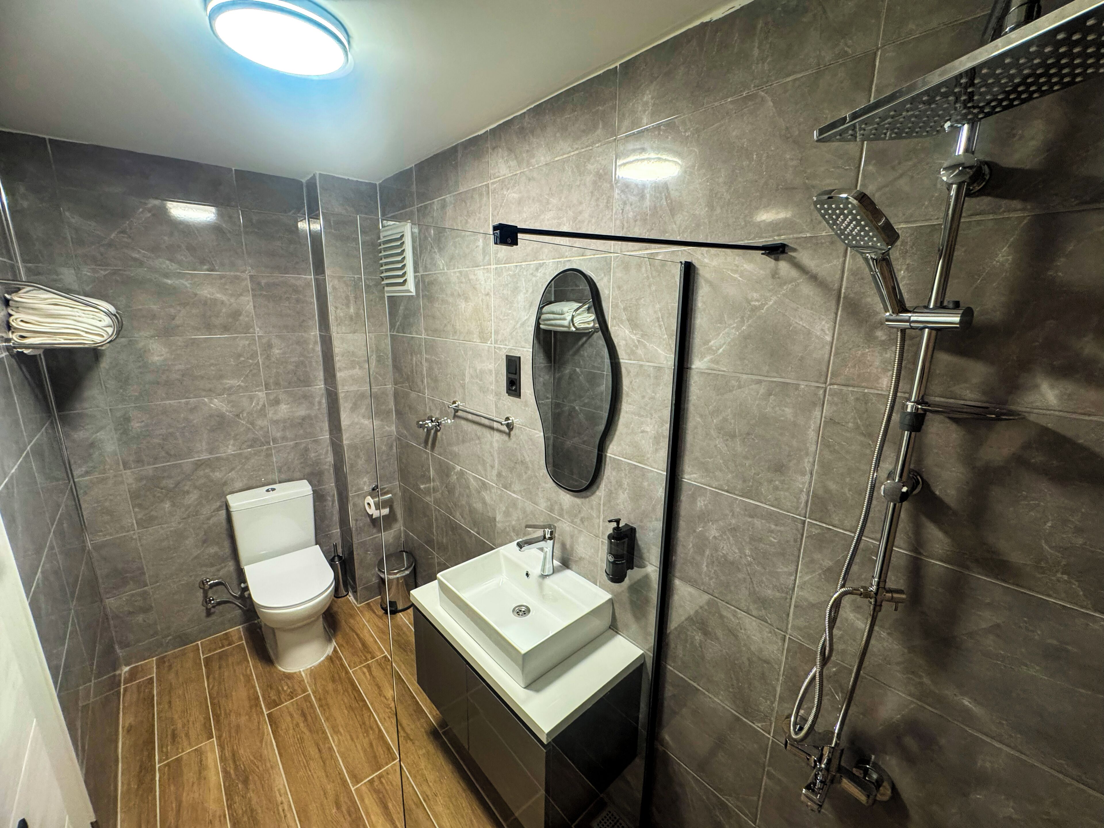 Un baño moderno con inodoro, lavabo, espejo, toallero, ducha y una luz en el techo.
