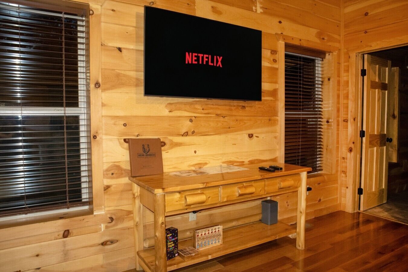 Un intérieur en bois avec un écran de télévision fixé au mur affichant le logo « NETFLIX », un bureau en bois avec des tiroirs et une porte sur la droite.