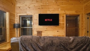 Una habitación acogedora con un sofá de cuero, una manta, un televisor de pantalla plana montado en una pared de madera y una ventana con persianas.