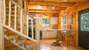 Interior de una cabaña de madera con cocina, comedor y una escalera.
