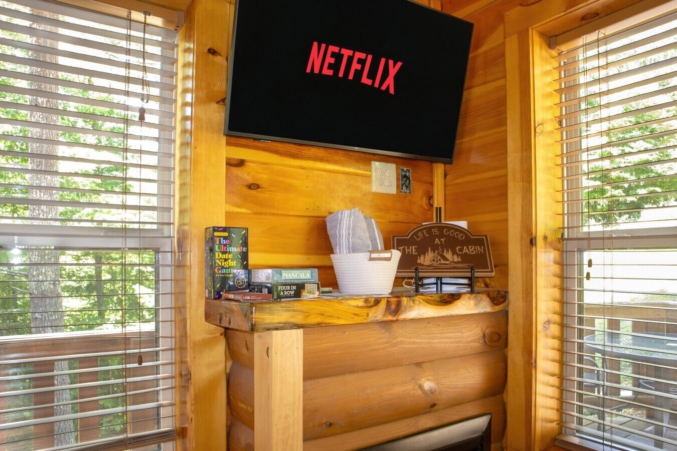 Interior de uma cabana de madeira com uma televisão presa na parede exibindo 'NETFLIX', uma estante de madeira com jogos de tabuleiro e uma placa com a inscrição 'LIFE IS GOOD AT THE CABIN'.