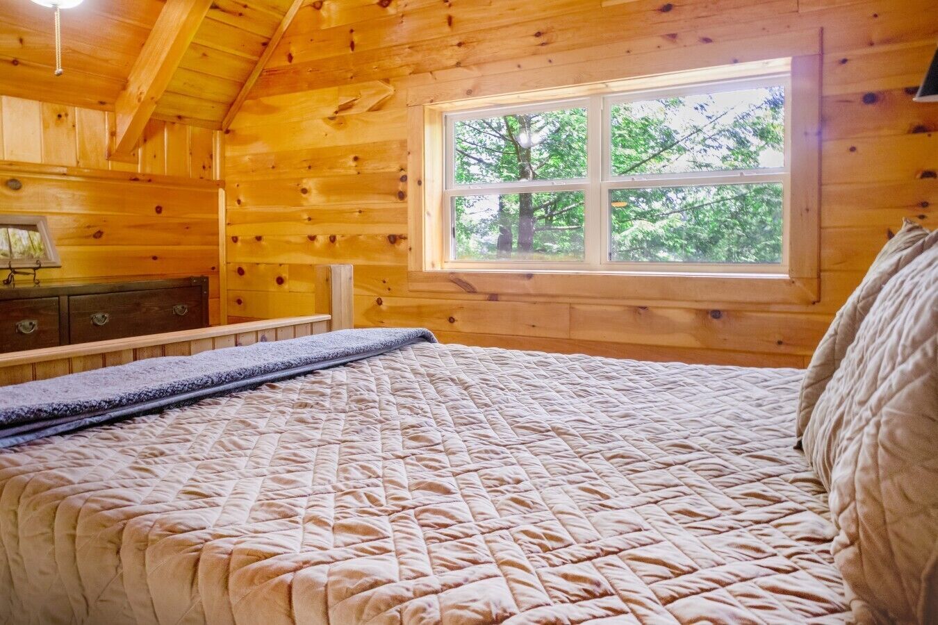 A wooden cabin bedroom with a bed, a window with a view of trees, and a dresser.