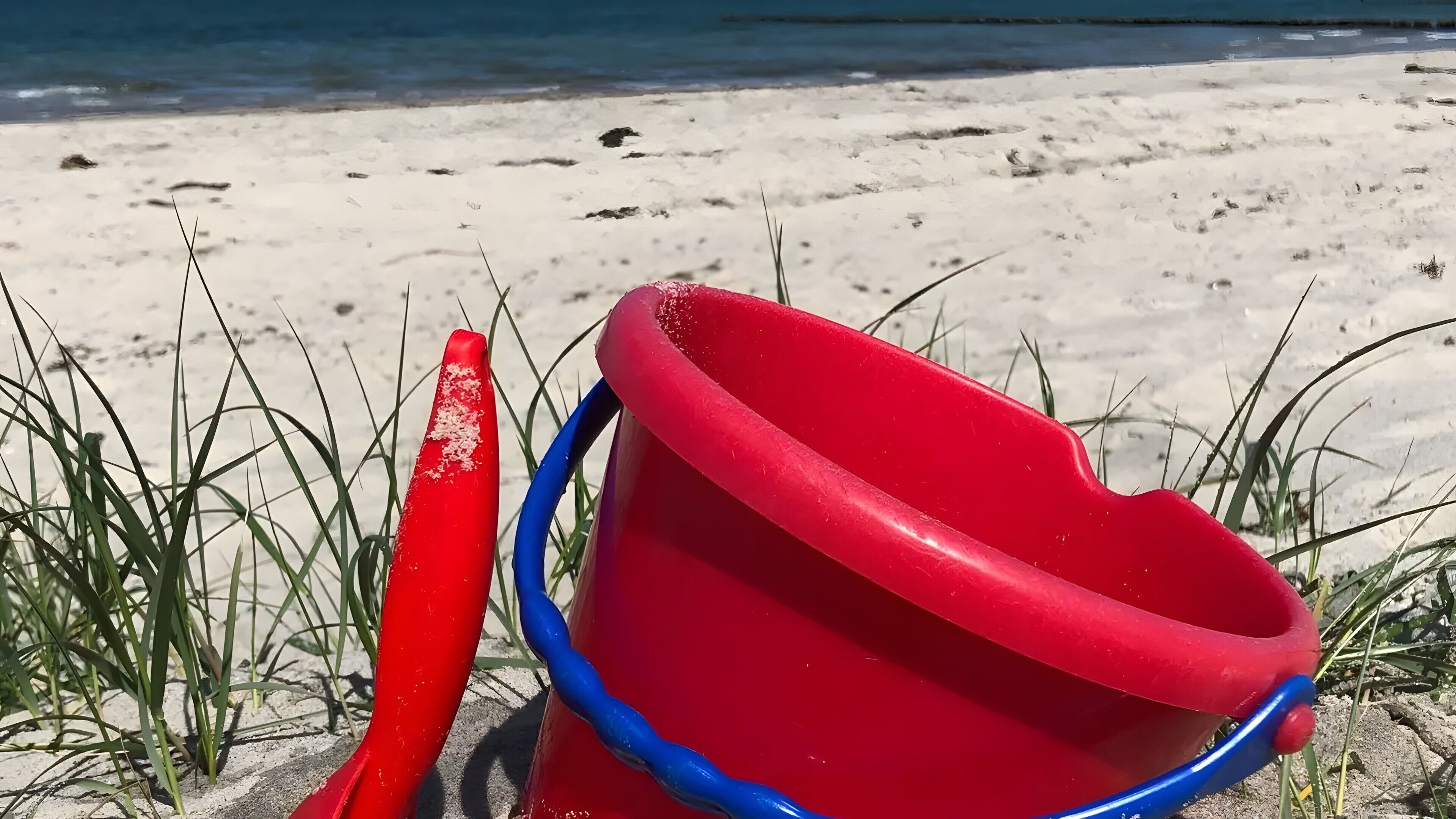Un juguete de playa rojo y azul sobre la arena, con césped al fondo.