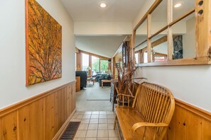 A hallway with wooden paneling, a bench, a large abstract painting, and a view into a living area with a couch and a dining table.