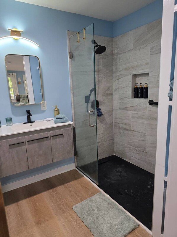 A modern bathroom with a glass shower enclosure, a vanity with a mirror, and a small shelf with toiletries. - Modern Guesthouse Near Beaches & FLETC (Brunswick)