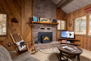 Ein gemütliches Wohnzimmer mit Kamin aus Stein, Holzwänden, einem Fernseher und einem Schaukelstuhl.