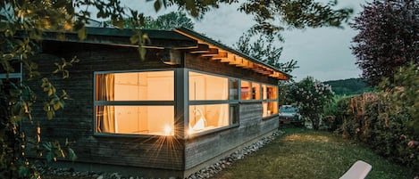 Une maison moderne dotée de grandes fenêtres, d’une façade en pierre et d’un jardin agrémenté d’un fauteuil de relaxation.