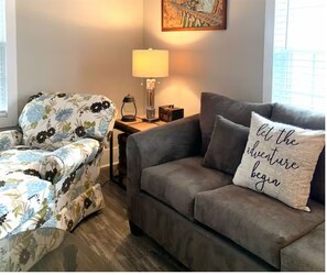 Ein Wohnzimmer mit einem floralen Sessel, einem grauen Sofa, einer Lampe und einem Kissen mit dem Aufdruck 'let the adventure begin'.