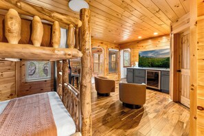 Ein Holzblockhaus-Interieur mit einem Bett, zwei Sesseln, einer Küchenzeile und einem großen Fenster mit Blick.
