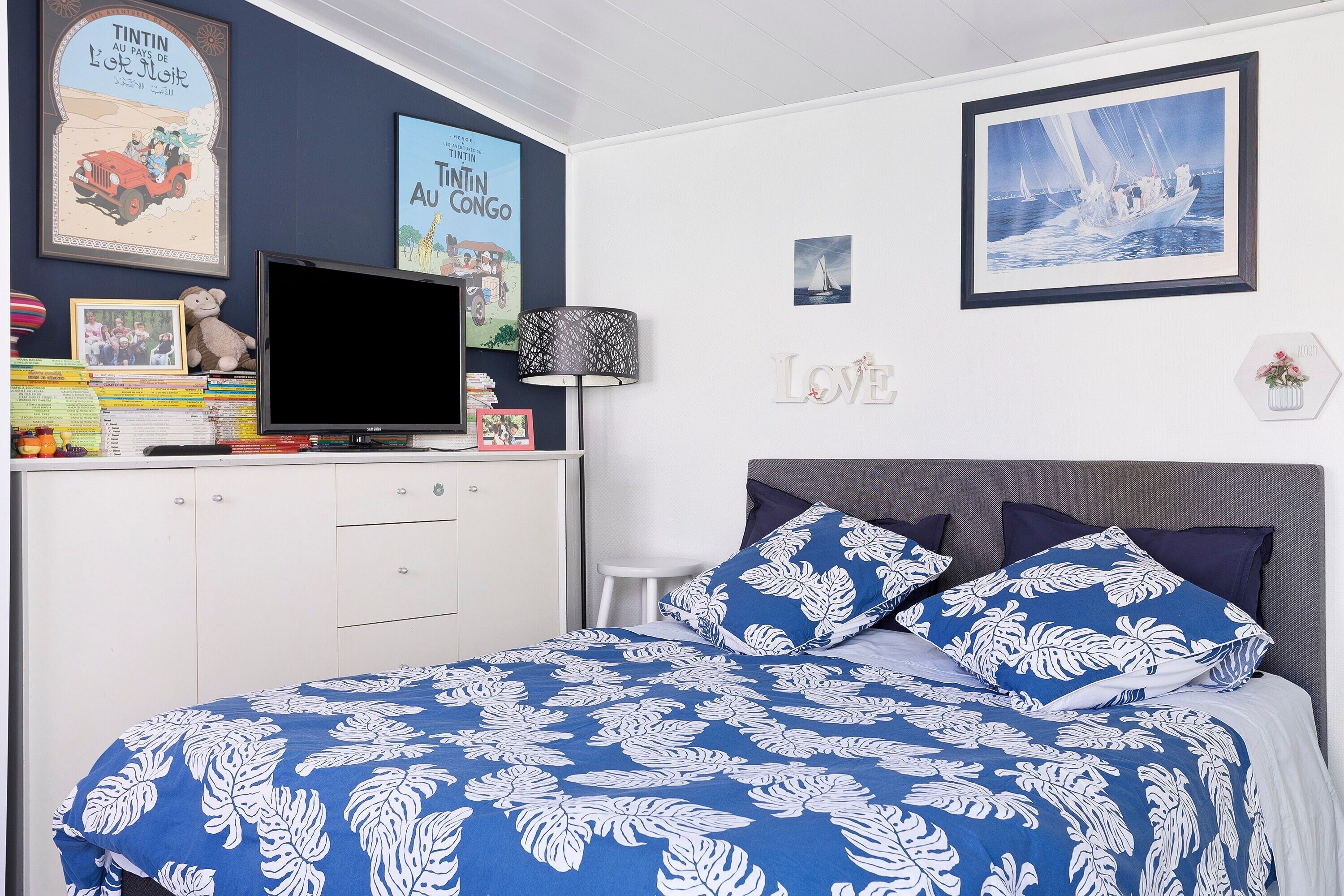 Ein Schlafzimmer mit einem Bett, einem Fernseher, einem Schreibtisch mit Büchern und einer Lampe sowie Wanddekorationen, darunter gerahmte Bilder und das Wort 'LIEBE'.