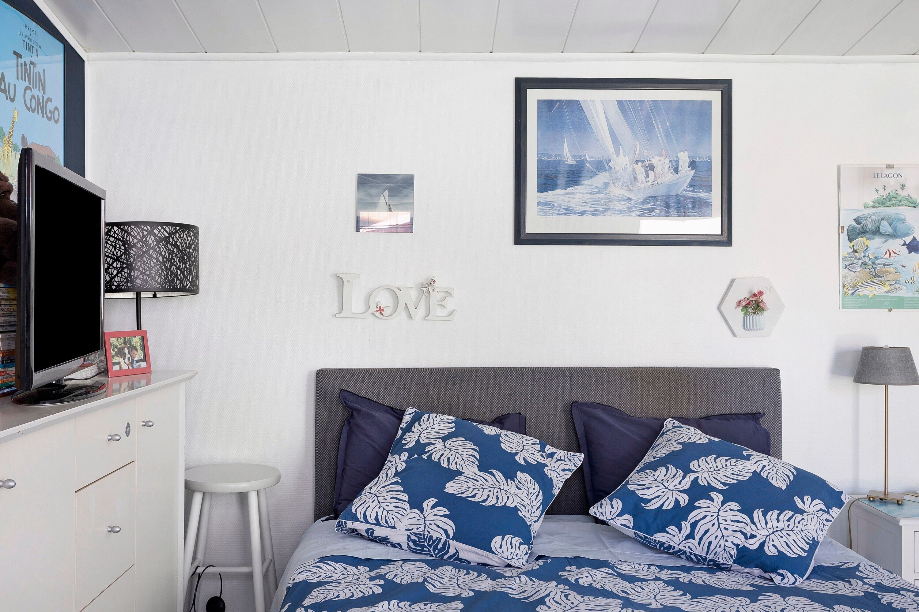 Een slaapkamer met een bed, een televisie, een witte ladekast, een klein krukje en wanddecoraties, waaronder een ingelijste foto en het woord 'LOVE'.