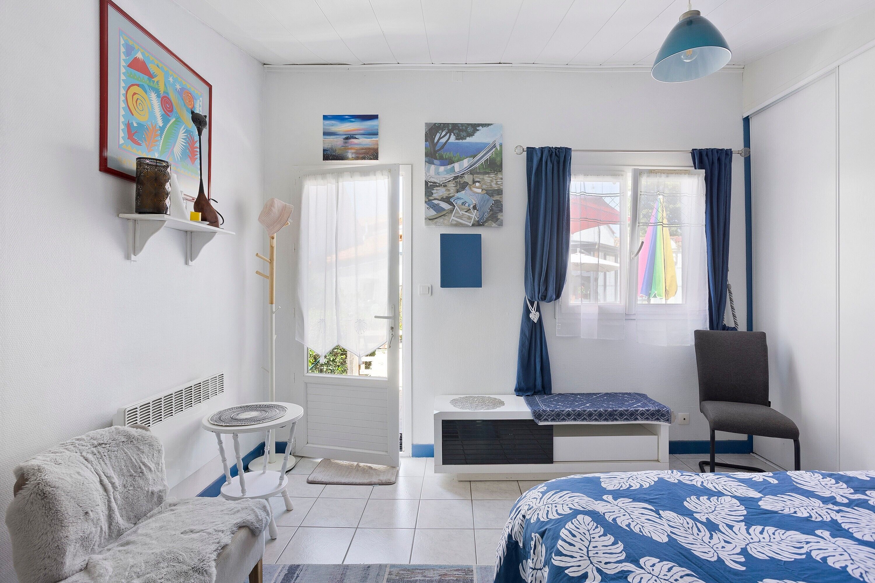 Una habitación con cama, una silla, una mesita, una estantería con adornos y una ventana con cortinas.
