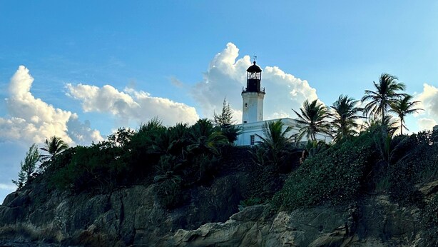 A lighthouse on a rocky cliff with waves crashing below, palm trees, and a clear sky. - TheTimeless Haven PR@ Oceanfront Escape- Maunabo (Maunabo)