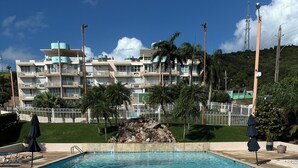 A clear swimming pool with a metal ladder, surrounded by a concrete deck. In the background, there is a multi-story building with balconies and a white fence. - TheTimeless Haven PR@ Oceanfront Escape- Maunabo (Maunabo)