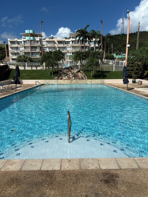 A clear swimming pool with a metal ladder, surrounded by a concrete deck. In the background, there is a multi-story building with balconies and a white fence. - TheTimeless Haven PR@ Oceanfront Escape- Maunabo (Maunabo)