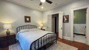 A bedroom with a bed, bedside lamps, a nightstand, a door, and a bathroom visible through an open door. - Charming, Spacious Victorian Near Downtown Dallas (Dallas)