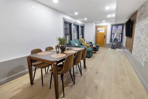 A dining area with a wooden table and chairs, a sofa, and a TV mounted on a brick wall. - North End 2BR Home / Prime Boston Location (Boston)