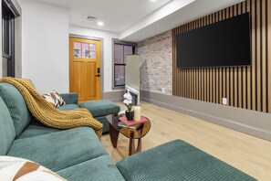 A living room with a sofa, a coffee table, a TV mounted on the wall, and a wooden door. - North End 2BR Home / Prime Boston Location (Boston)