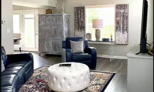Ein Wohnzimmer mit blauem Sessel, weißem Hocker, einem dunkelbraunen Ledersofa und einem Teppich mit Muster.